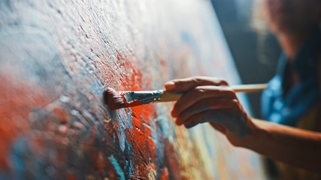 Female artist works on abstract oil painting, moving paint brush energetically she creates modern masterpiece. dark creative studio where large canvas stands on easel illuminated. low angle close up