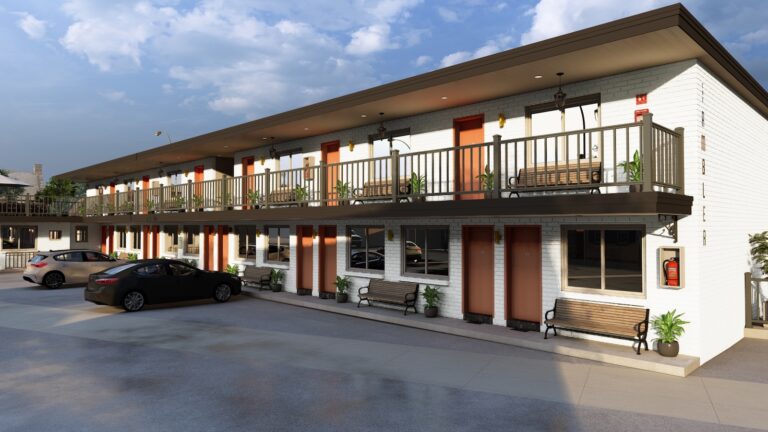 A two-story motel with white brick walls, red doors, metal railings, parked cars in front, and potted plants along the walkway on a sunny day.