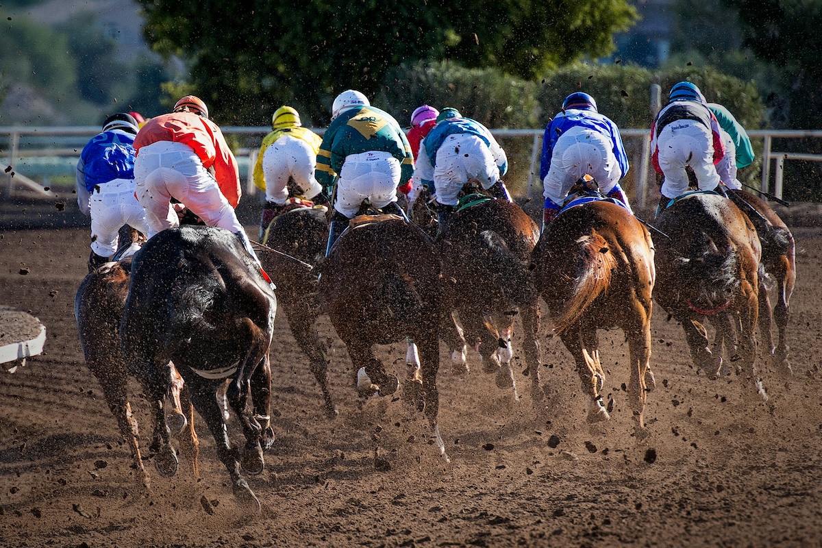 Jockeys in colorful uniforms ride horses at full speed on a dirt racetrack, with dust and debris flying up behind them.