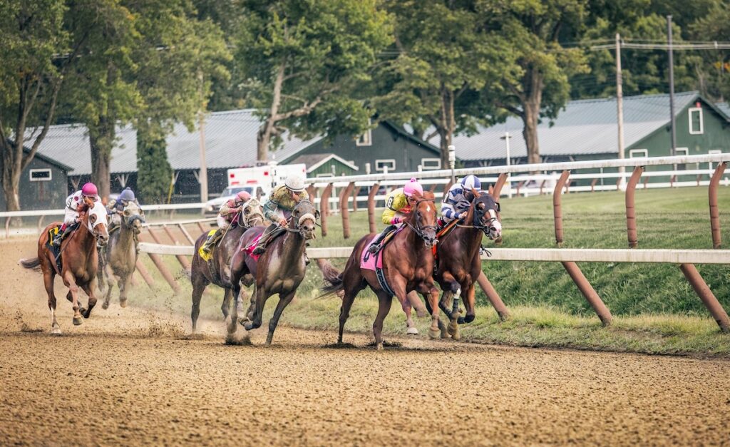Several jockeys ride horses around a dirt racetrack, with trees and green barns in the background.