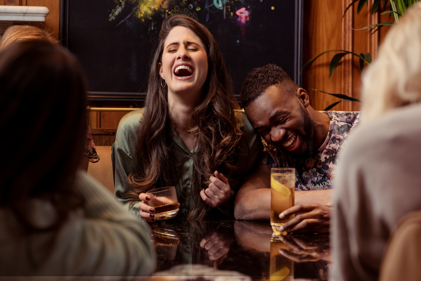 A woman and a man sit at a table, laughing and holding drinks, with two other people partially visible in the foreground.