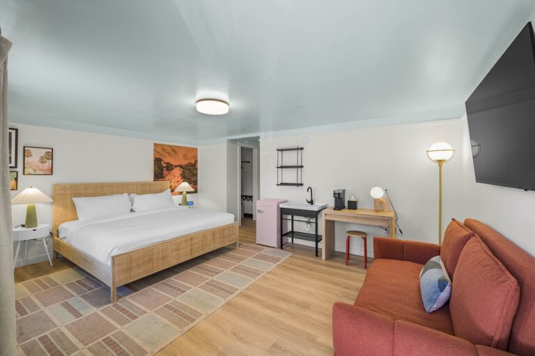 A modern hotel room with a king bed, red sofa, desk, wall-mounted TV, kitchenette with a pink fridge, and a large area rug on wooden flooring.