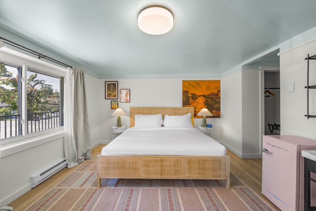 A bright bedroom with a large bed, two nightstands with lamps, a pastel rug, wall art, an open closet, a pink mini fridge, and a window with curtains.