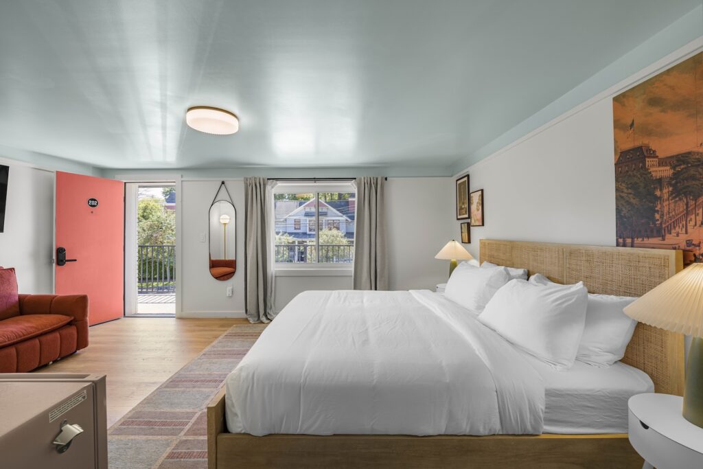 Bright hotel room with a king bed, red sofa, wall art, mirror, and a door leading to an outdoor balcony with a view of houses.