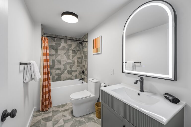 Modern bathroom with patterned tile floor and shower wall, orange checkered shower curtain, white toilet, gray vanity with illuminated mirror, and a wall towel rack.