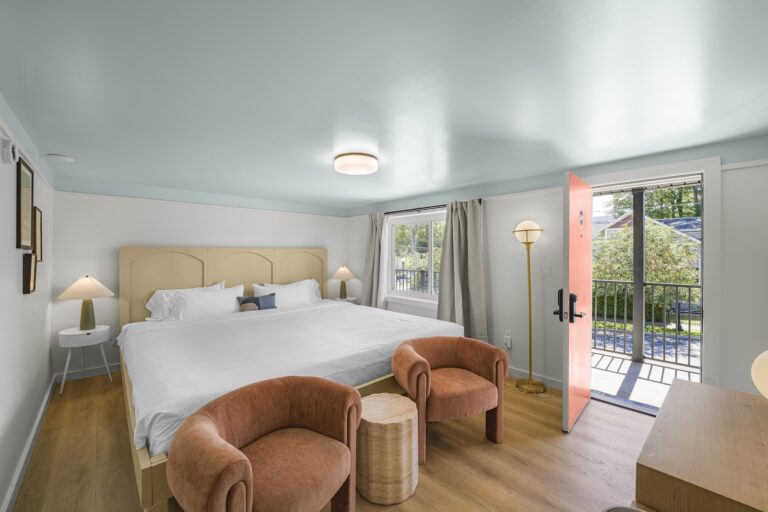 A bedroom with a large bed, two brown armchairs, a small wooden table, side tables with lamps, and an open door leading to a balcony. Natural light fills the room.