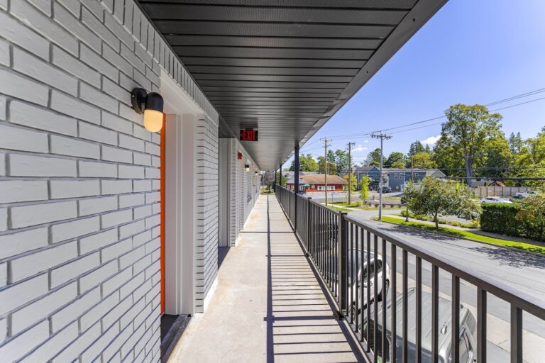 A long, outdoor hallway with white brick walls, black railing, and overhead lights, overlooking a street with trees and parked cars on a sunny day.