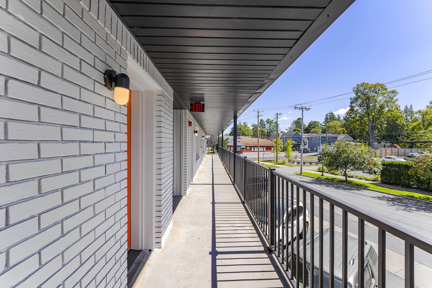 A long, outdoor hallway with white brick walls, black railing, and overhead lights, overlooking a street with trees and parked cars on a sunny day.