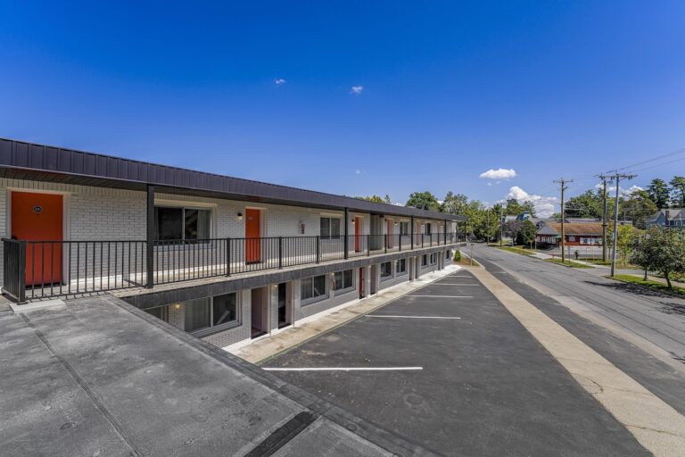 Two-story motel with red doors, exterior corridors, and an empty parking lot, located next to a quiet street under a clear blue sky.