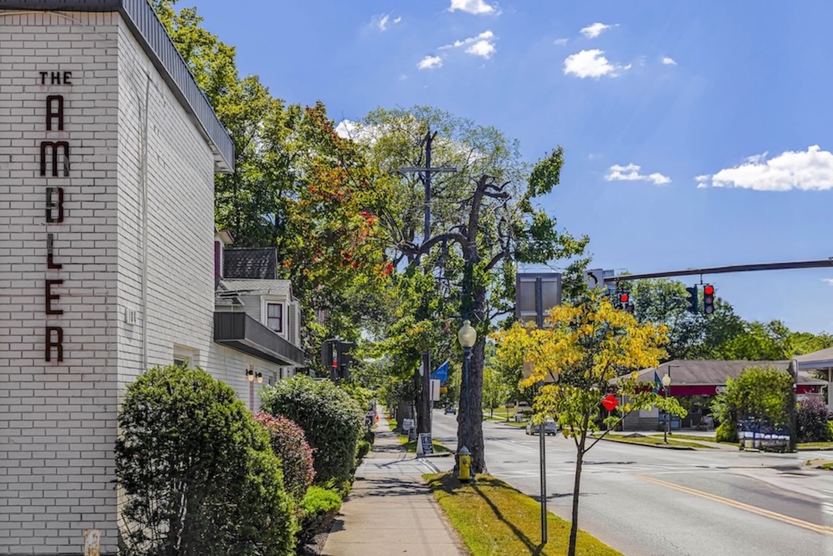 A street view shows a building with “The Ambler” sign, trees, shrubbery, a sidewalk, and a traffic light in the background under a clear blue sky.
