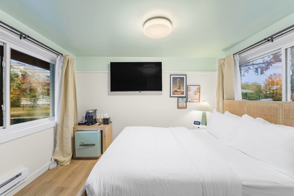A tidy bedroom with a large bed, wall-mounted TV, framed pictures, a minibar, coffee maker, and windows with beige curtains showing trees outside.