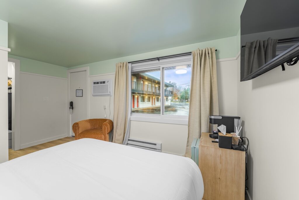 A motel room with a bed, orange armchair, small dresser with a coffee maker, wall-mounted TV, and a window showing an outdoor view of the motel exterior.