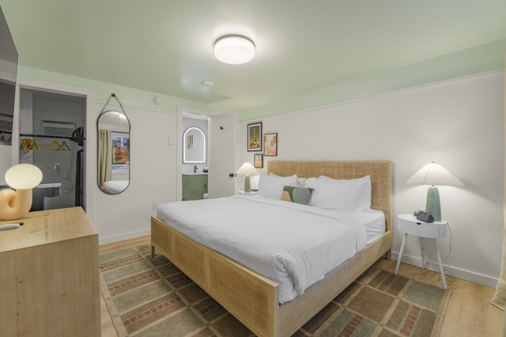 A bedroom with a large bed, two lamps on bedside tables, a dresser with a lamp, a wall mirror, and a view into an adjacent bathroom.