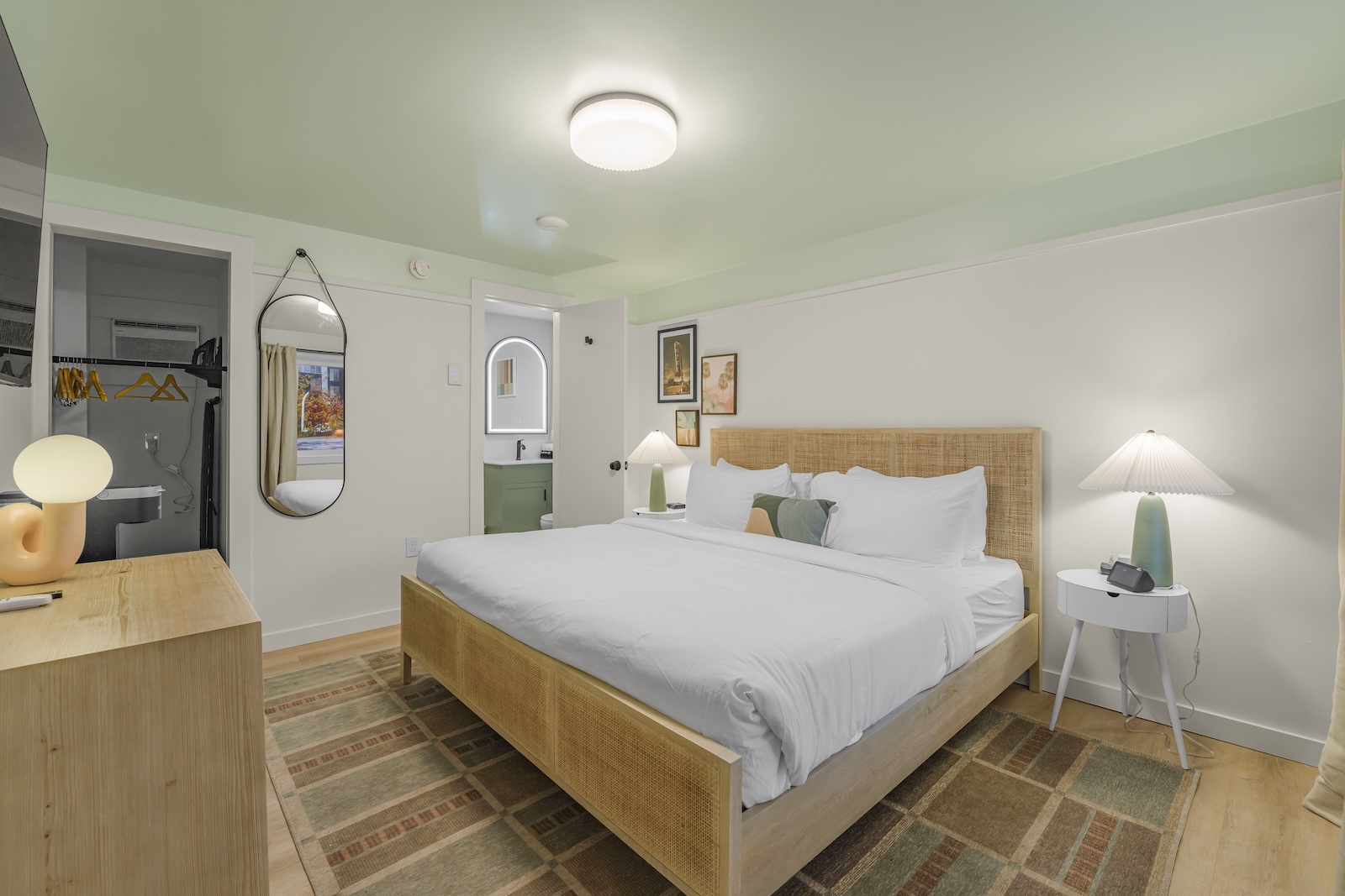 A bedroom with a large bed, two lamps on bedside tables, a dresser with a lamp, a wall mirror, and a view into an adjacent bathroom.