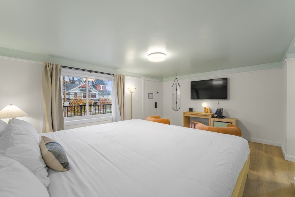 Bright bedroom with a large bed, TV, desk with two chairs, lamp, and window overlooking houses and trees. The walls and ceiling are painted light colors.