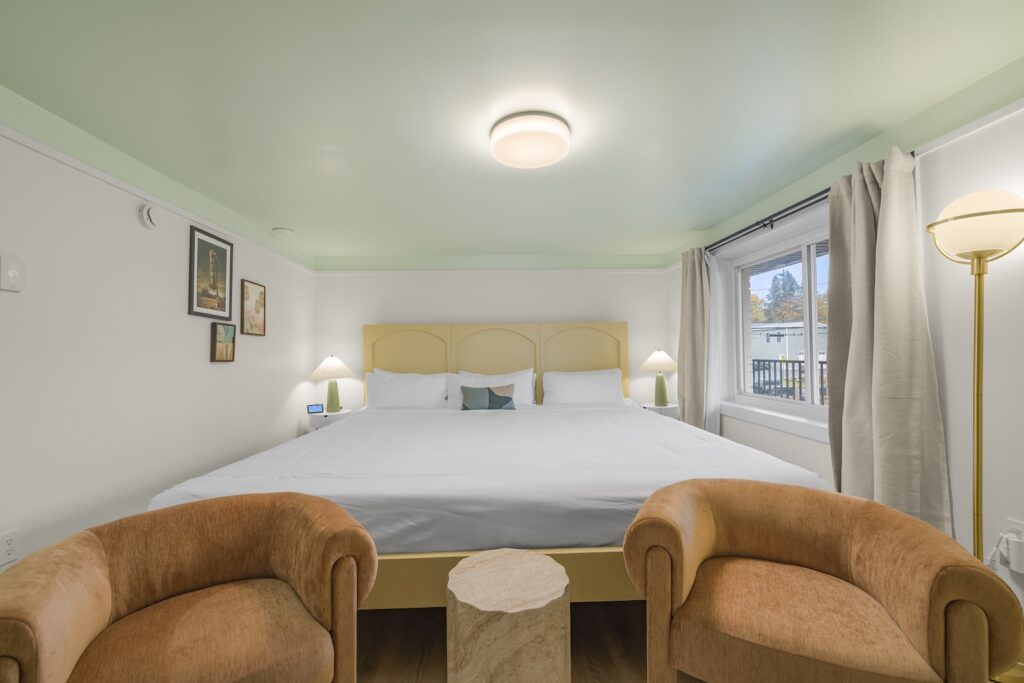 A neatly made bed with white linens is centered between two nightstands, flanked by tan armchairs and a round table in a well-lit room with large window and wall art.