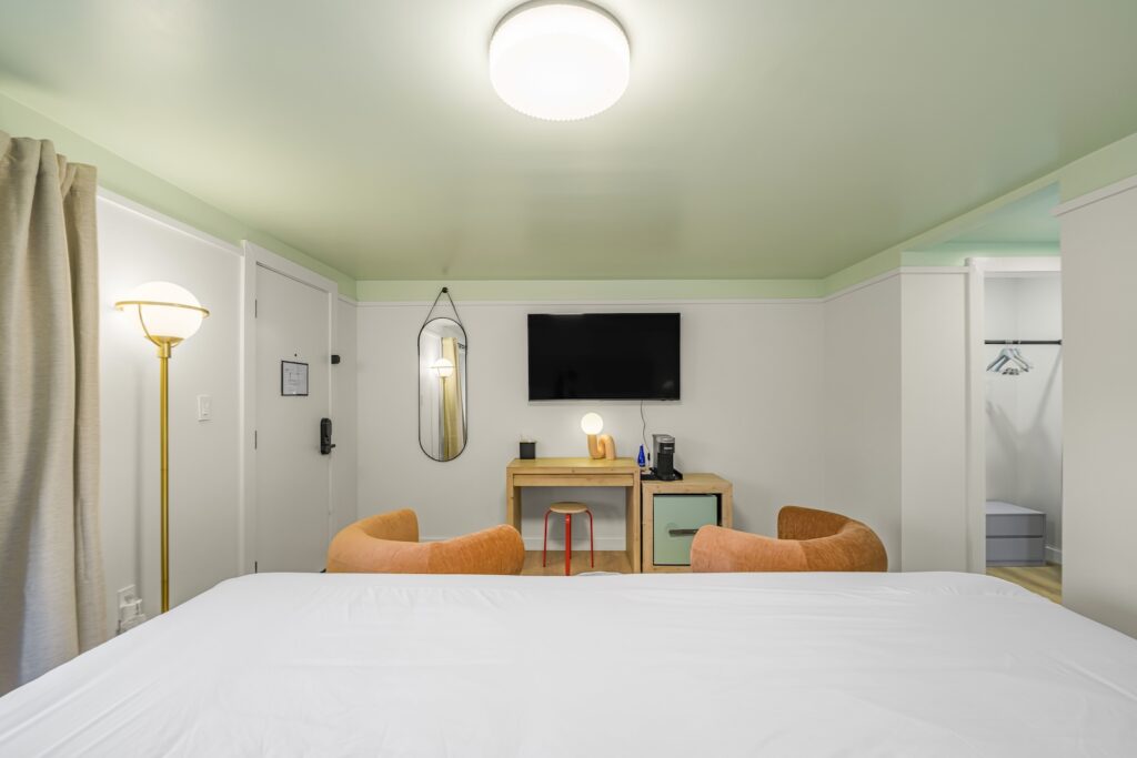 Modern hotel room with a neatly made bed, two orange chairs, a desk with a lamp, wall-mounted TV, and an open closet in the background.